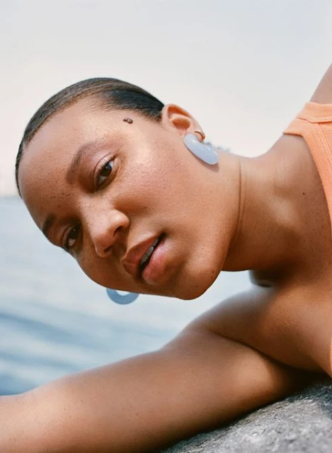 Close-up of a young African-American woman wearing hoop earrings, reclining by the water, and smiling in natural daylight.
