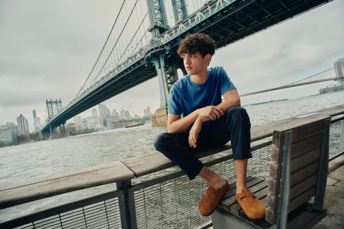 Man sits on a waterfront bench with the Manhattan Bridge in the background, wearing a blue t-shirt and brown slip-on shoes.