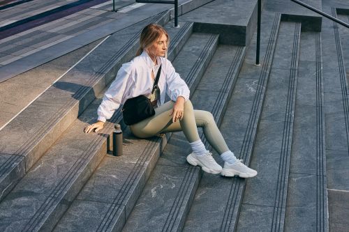 Athletic woman in white jacket and olive leggings sits on gray steps with water bottle and black bag in an urban setting.
