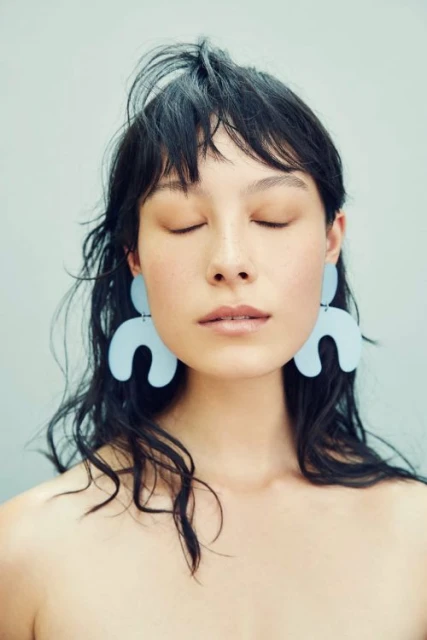 Portrait of a woman with eyes closed wearing large, modern earrings, captured in soft lighting against a muted pastel backdrop.