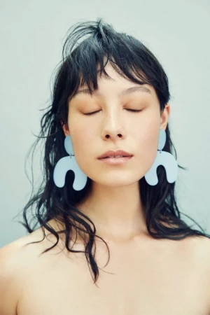 Portrait of a woman with eyes closed wearing large, modern earrings, captured in soft lighting against a muted pastel backdrop.