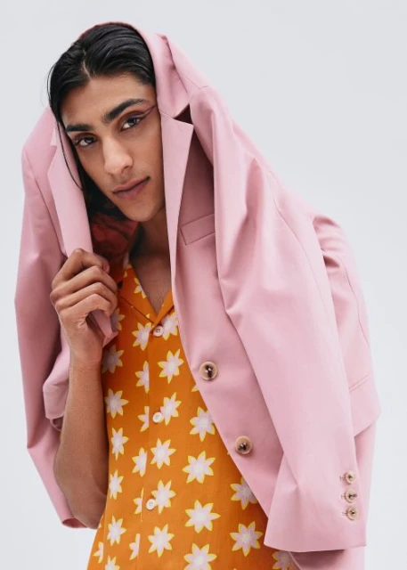 Stylish man with pink blazer over his head poses in bright orange shirt with floral pattern in front of a plain white background.
