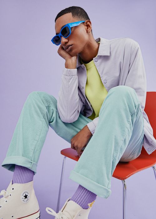 Man in retro eyewear and pastel corduroy seated on orange chair in a studio with lavender background wearing cream high-top sneakers.
