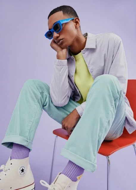 Man in retro eyewear and pastel corduroy seated on orange chair in a studio with lavender background wearing cream high-top sneakers.