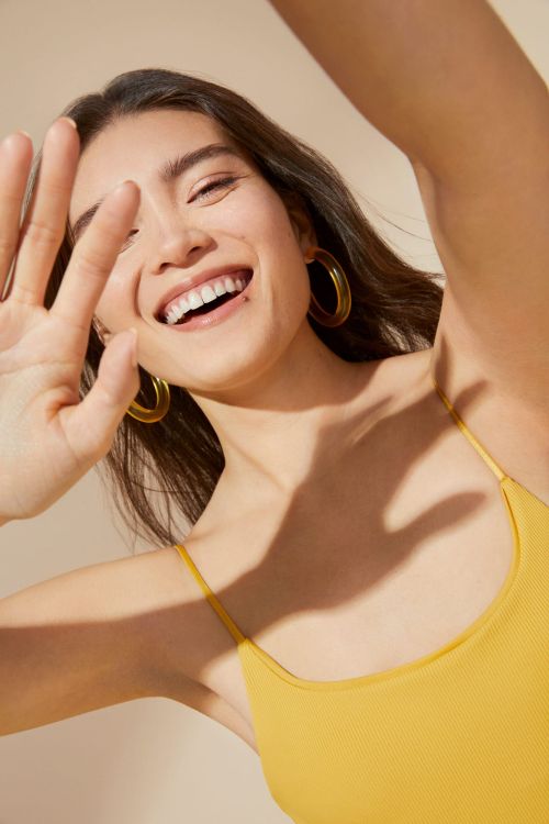 Smiling woman with raised hand and hoop earrings wearing yellow tank top against a neutral background in a portrait shot.