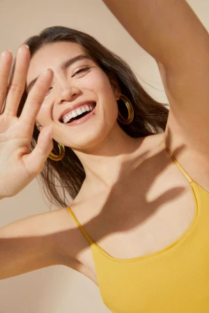 Smiling woman with raised hand and hoop earrings wearing yellow tank top against a neutral background in a portrait shot.