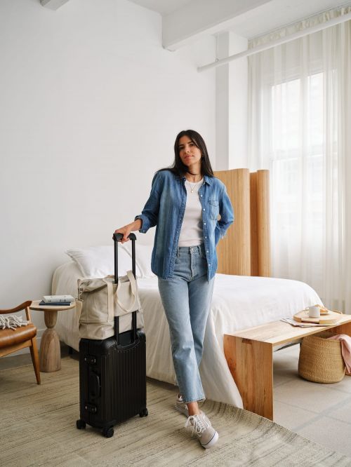 Young woman posing indoors with a black suitcase and tote bag in a bright, neutral-toned room, wearing a denim shirt and jeans.