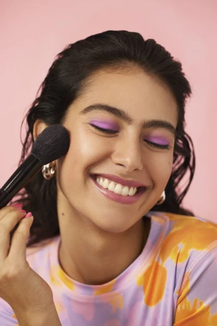 Smiling woman applying makeup with a brush in a beauty shot against a light pink background wearing a patterned shirt and gold earrings.