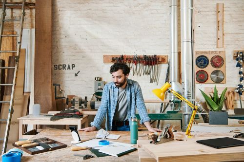 Carpenter works in a wood shop with hand tools, power tools, and lumber in a cluttered workshop with a yellow desk lamp.