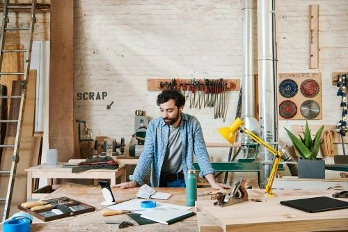 Carpenter works in a wood shop with hand tools, power tools, and lumber in a cluttered workshop with a yellow desk lamp.
