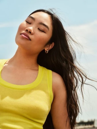 Close-up portrait of an East Asian woman with dark hair wearing a bright yellow tank top against a pale blue sky background.