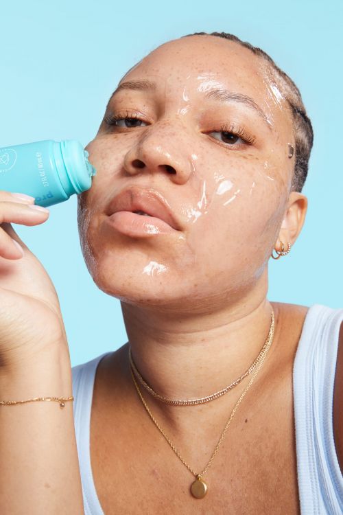 Adult woman with skincare on face applying cosmetic product from blue tube. Close-up beauty shot against light blue backdrop.