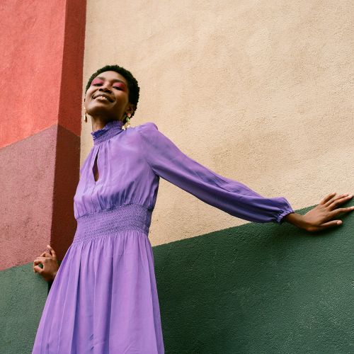 A woman in a long-sleeve purple dress leans against a multicoloured wall, smiling with visible red eyeshadow and short dark hair.