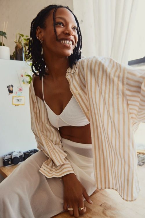 Smiling African-American woman in a striped shirt and white pants sits on a wooden surface with cameras and plants in the background.