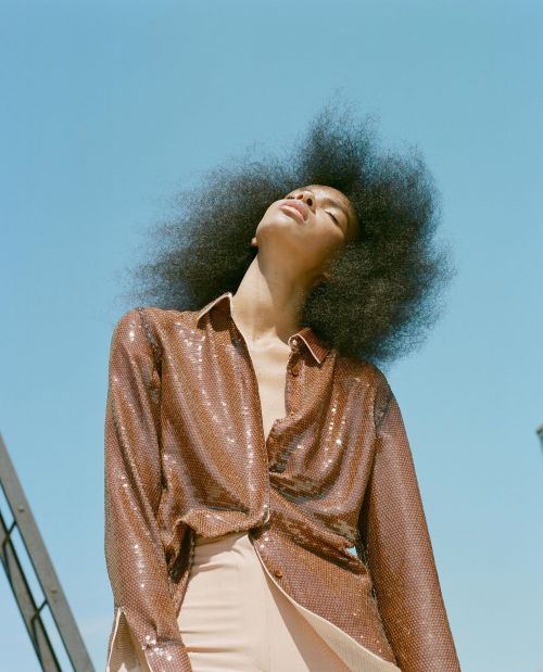 Woman with an afro poses in front of a blue sky wearing a sparkling brown shirt and beige pants in an outdoor fashion shot.