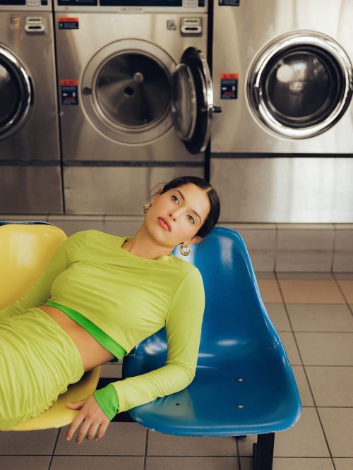Woman in green loungewear poses on colorful seats at a laundromat, with stainless steel washing machines in the background.