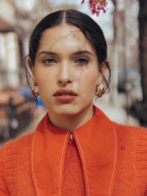 Portrait of a young woman with gold hoop earrings, wearing an orange top with a white collar, standing outdoors near flowering tree.