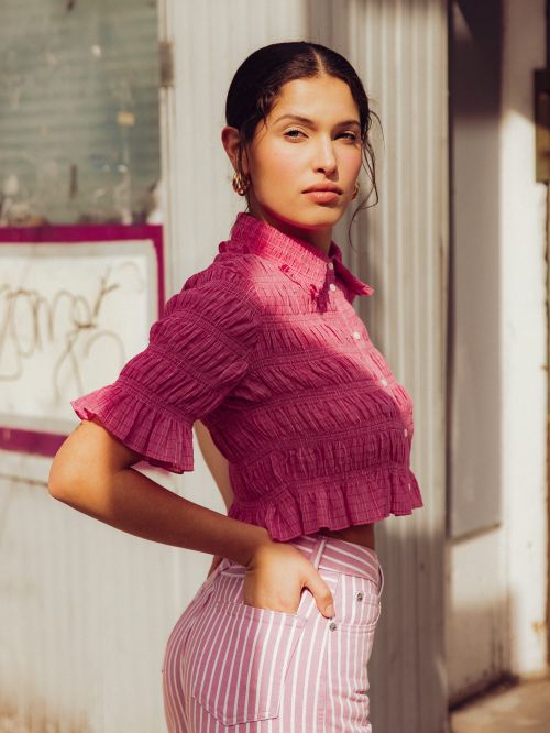 Portrait of a young woman in a pink crop top and striped pants, posing in front of a building on a sunny day.