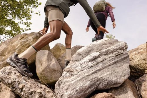 Two hikers traverse a rocky landscape. One wears a jacket, shorts, socks, and trail shoes, while the other has a backpack and climbs a rock.