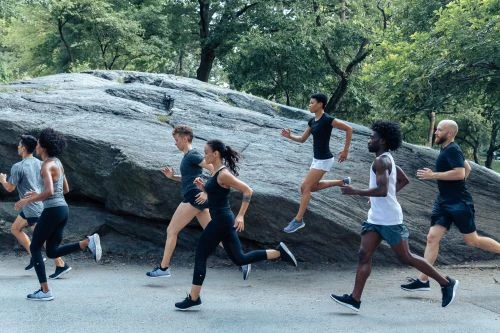 Group of diverse adults runs on path in park, wearing athletic clothes and shoes; trees and gray rocks in background on overcast day.