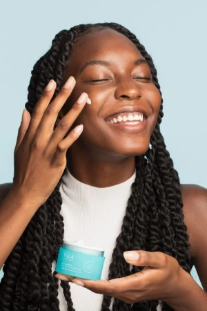Smiling woman applying cream to face. Focus on skincare routine with Tula product, blue background, showcasing beauty and self-care.