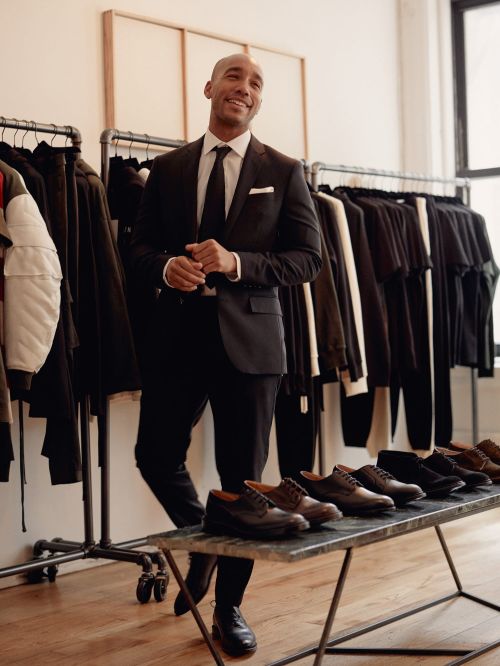 Man in a dark suit stands next to a table displaying dress shoes, with a rack of clothing in the background, in a neutral indoor setting.