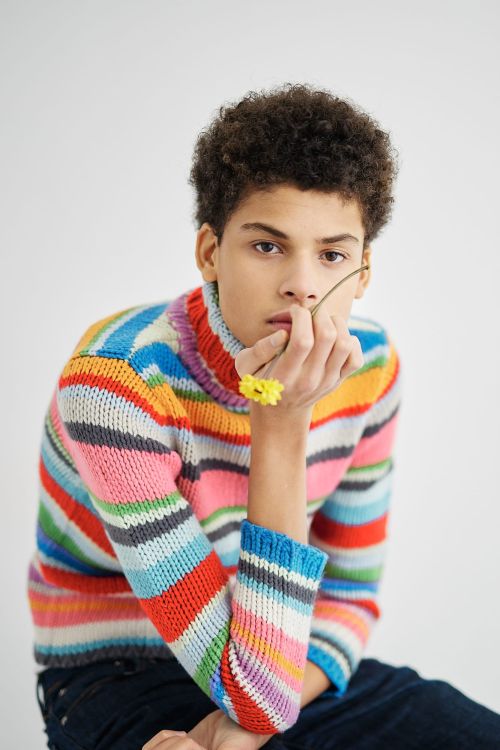 Young man with curly hair wearing a colorful striped turtleneck sweater holding small yellow flowers in front of a white background.