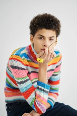 Young man with curly hair wearing a colorful striped turtleneck sweater holding small yellow flowers in front of a white background.