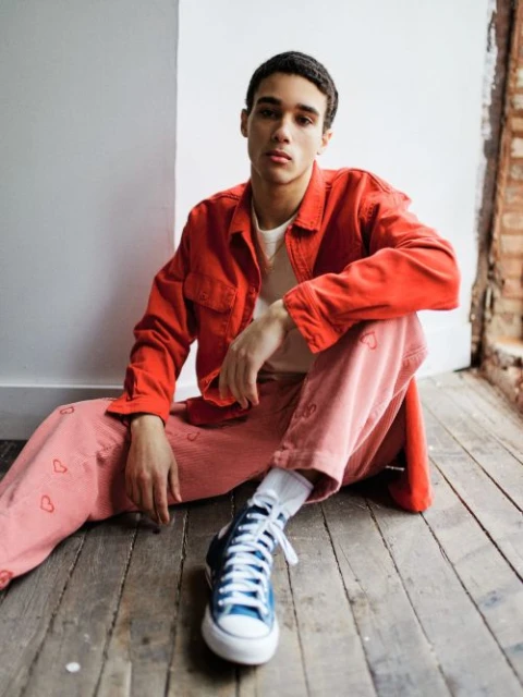 A young man in a red jacket and pink pants with heart details poses indoors against a white and brick wall while sitting on wood floors.