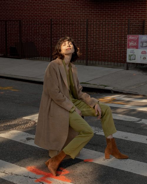 A woman in a tan coat sits on a crosswalk with a brick wall backdrop, basking in sunlight, showcasing fashion and urban style.