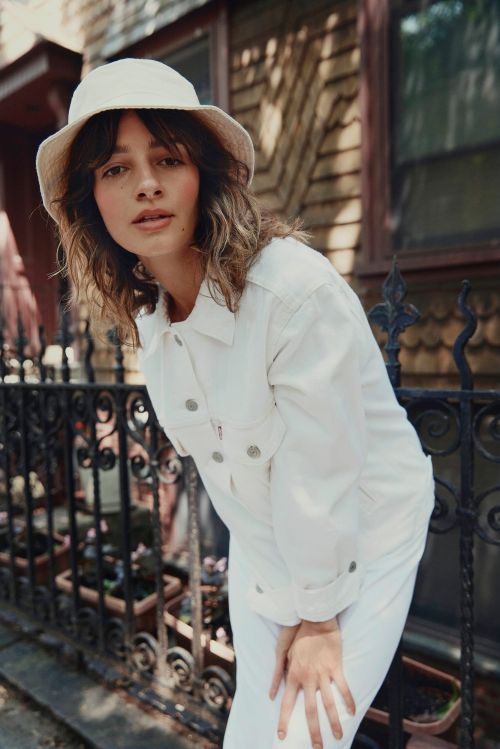 Portrait of a young woman in white denim jacket and hat posing outdoors near a dark iron fence and house with brown wood siding.