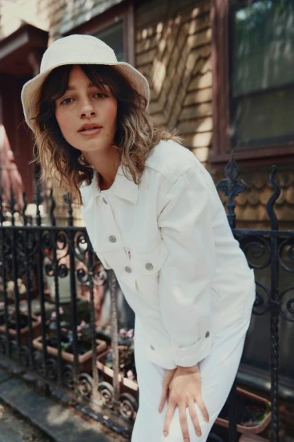 Portrait of a young woman in white denim jacket and hat posing outdoors near a dark iron fence and house with brown wood siding.