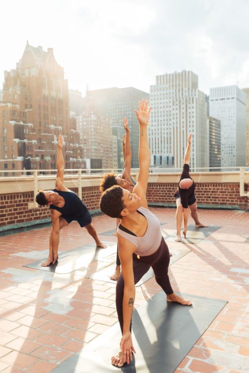 Group of adults in yoga poses on a rooftop against city buildings in daylight, performing yoga on mats wearing activewear.