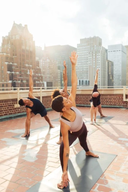 Group of adults in yoga poses on a rooftop against city buildings in daylight, performing yoga on mats wearing activewear.