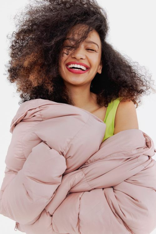 Smiling woman with curly hair and bright red lipstick wearing a lime green top and holding a puffy pink jacket against a white background.