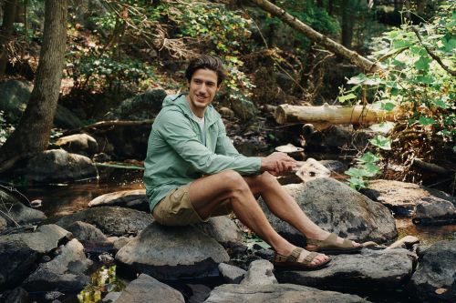 Man in light green jacket and shorts sits on rocks in a forest stream, smiling at the camera in a medium shot on a sunny day.