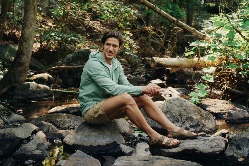Man in light green jacket and shorts sits on rocks in a forest stream, smiling at the camera in a medium shot on a sunny day.