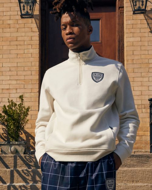 A young Black man poses on stone steps in a cream-colored pullover, plaid trousers, and neutral colors against a brick building backdrop.