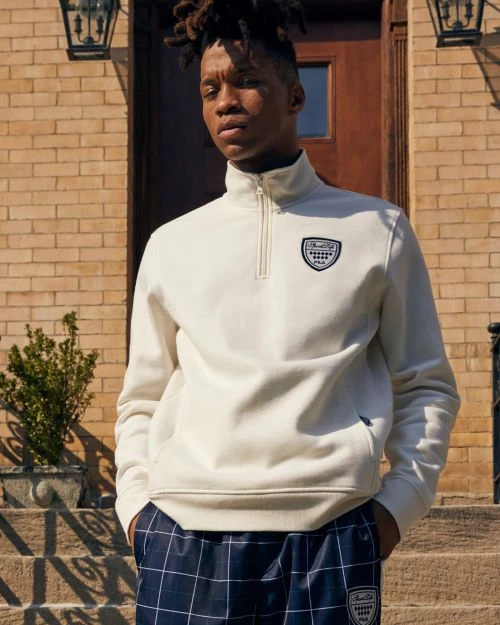 A young Black man poses on stone steps in a cream-colored pullover, plaid trousers, and neutral colors against a brick building backdrop.