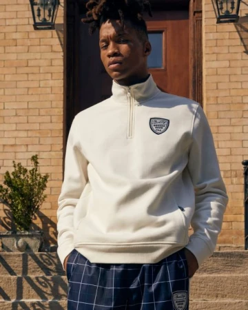 A young Black man poses on stone steps in a cream-colored pullover, plaid trousers, and neutral colors against a brick building backdrop.