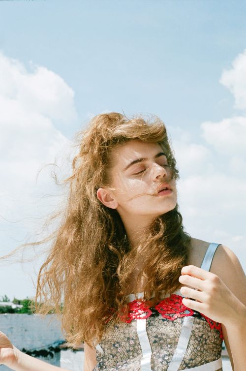 Portrait of a young woman with curly brown hair standing outside in a patterned top with red floral accents against a sunny, partly cloudy sky.