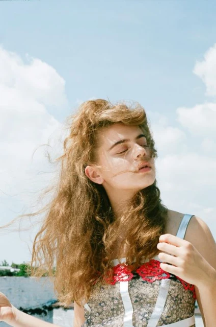 Portrait of a young woman with curly brown hair standing outside in a patterned top with red floral accents against a sunny, partly cloudy sky.