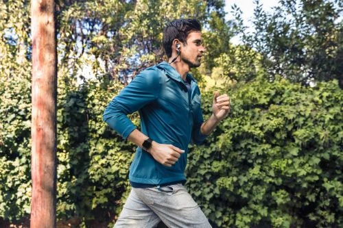 Adult male jogging outside wearing a blue jacket, gray sweatpants, headphones, and a watch, set against a green background.