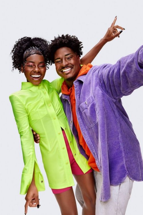Smiling man and woman pose for a selfie indoors. The woman wears a neon green shirt dress. The man has on a purple corduroy jacket.