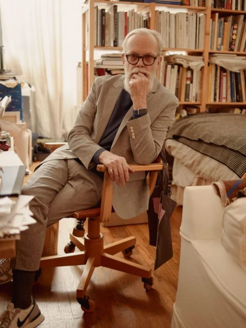 An older Caucasian man with a beard sits in a wooden chair, wearing a gray suit and glasses in a room filled with books and papers.