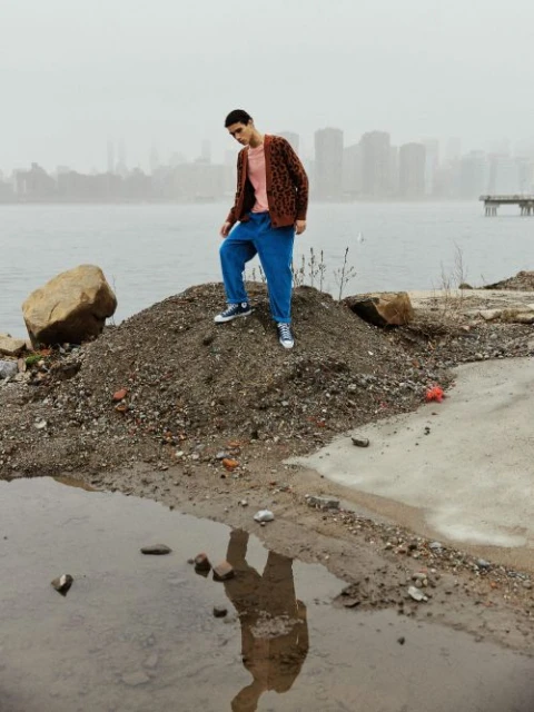 Young man in a cardigan and blue sweatpants stands on a dirt mound by the water, with city buildings visible in the distance.