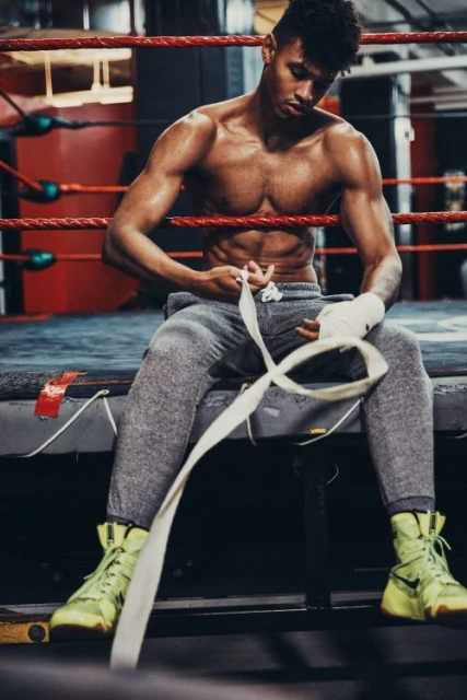 Shirtless man wraps his hands with boxing tape, sitting on the edge of a ring in a boxing gym with ropes and padded walls visible.