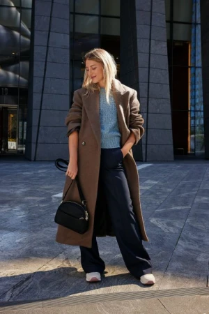 Woman wearing a long brown coat and light blue sweater holding a black handbag outdoors in New York