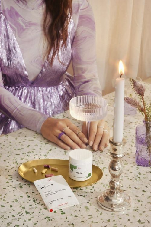 Woman sits at a table with a candle, beverage, and supplements, wearing a shiny lavender dress in an indoor setting.