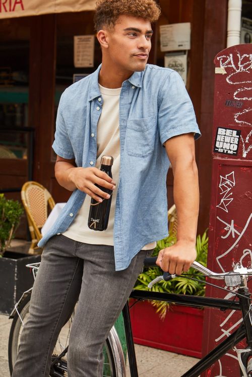 Young man in casual outfit standing with a bicycle outdoors, holding a water bottle in natural light, against urban backdrop.